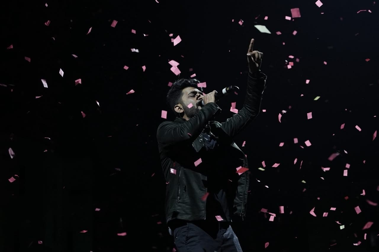 A singer mid-song with one arm raised as confetti falls through the stage light